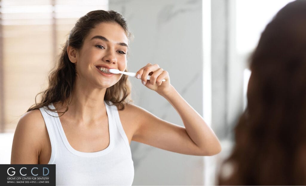 Smiling woman brushing her teeth in front of a bathroom mirror.