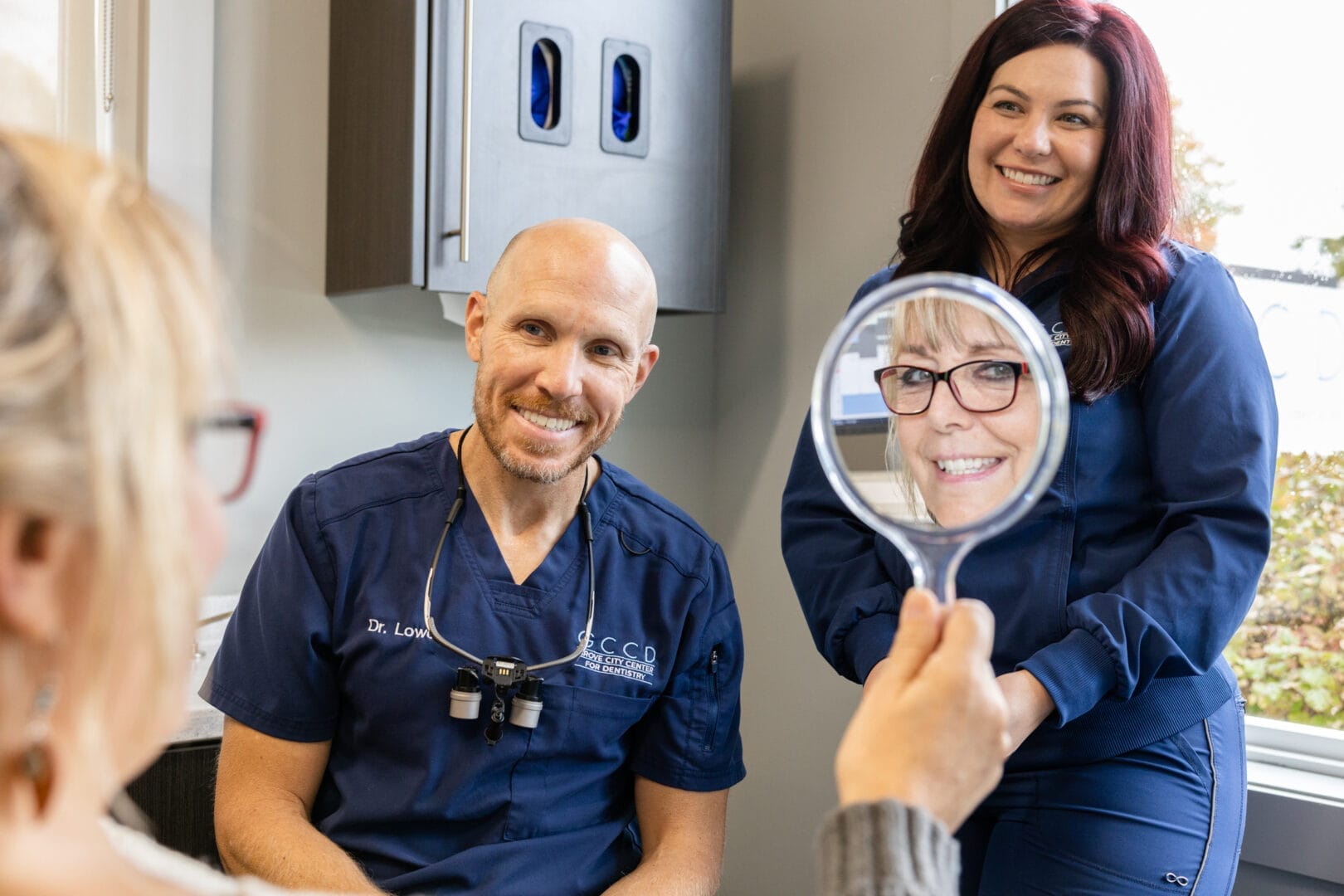 Dr. Kyle Lowe and dental hygienist smiling with patient looking in handheld mirror after checkup